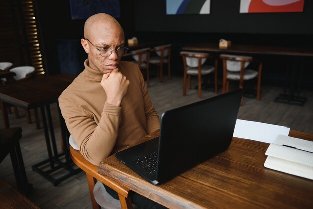 Óculos de estudante do sexo masculino negro sério tomando notas enquanto está sentado à mesa com o laptop e trabalhando em um projeto em um café aconchegante.