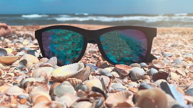Óculos de sol com lentes azuis ficam em uma praia de areia à beira-mar em um clima ensolarado. Conceito de recreação e turismo.