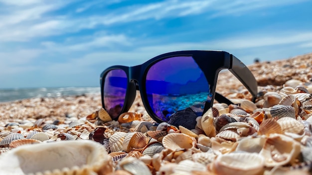 Óculos de sol com lentes azuis ficam em uma praia de areia à beira-mar em um clima ensolarado. Conceito de recreação e turismo.