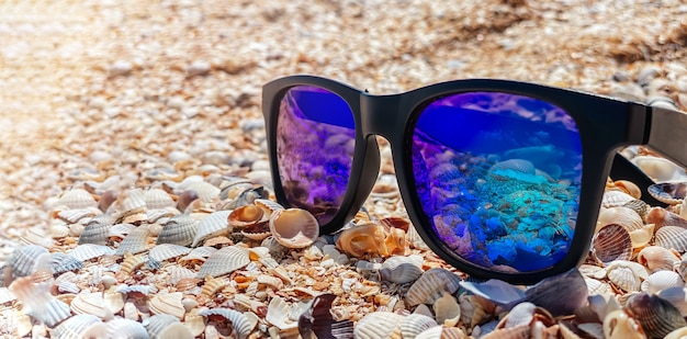 Óculos de sol com lentes azuis ficam em uma praia de areia à beira-mar em um clima ensolarado. Conceito de recreação e turismo.