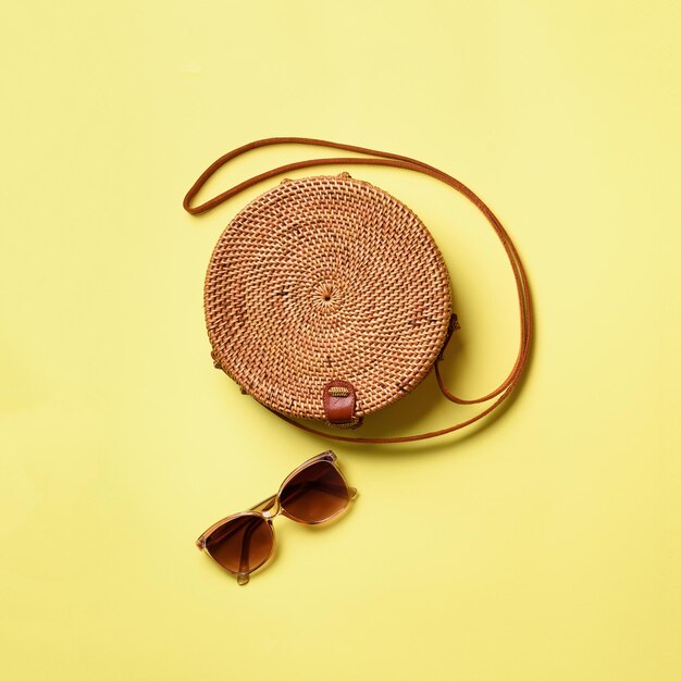 Óculos de sol de bolsa redonda de rattan em fundo amarelo Corte quadrado Vista superior com espaço para cópia Bolsa de bambu da moda e sapatos brancos Moda de verão plana lay Conceito de férias de viagem