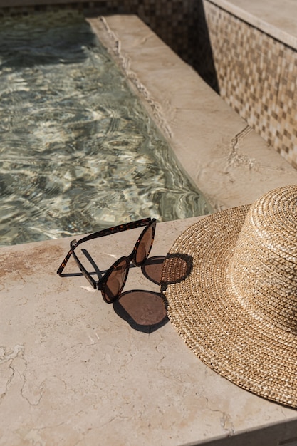 Óculos de sol e chapéu de palha ao lado da piscina de mármore com água azul clara com reflexos de sombra de luz solar de ondas