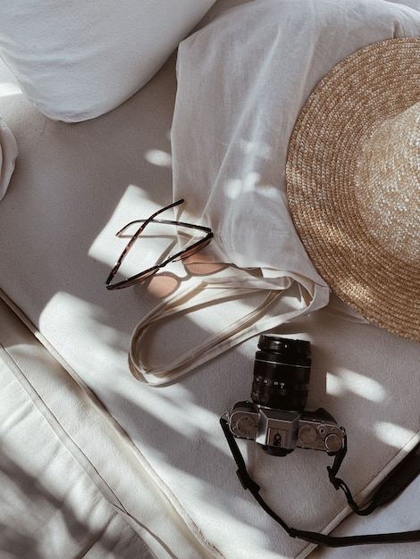Óculos de sol femininos, chapéu de palha, bolsa de compras, câmera fotográfica retrô em sofá branco com almofadas