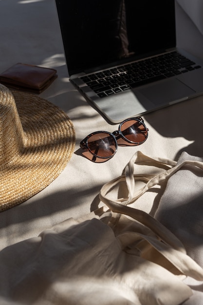 Óculos de sol femininos elegantes, chapéu de palha, bolsa de compras, laptop no sofá branco com almofadas.