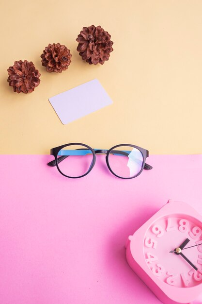 Óculos redondos na foto no estilo mínimo de verão em um fundo rosa pastel e amarelo. Despertador, flores de pinheiro, cartões de visita