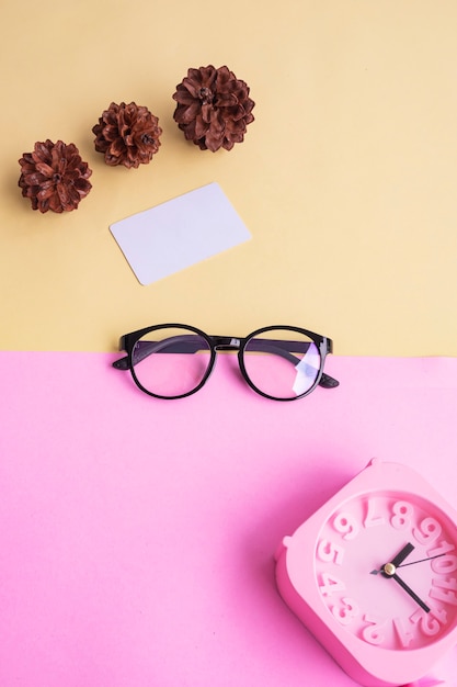 Óculos redondos na foto no estilo mínimo de verão em um fundo rosa pastel e amarelo. Despertador, flores de pinheiro, cartões de visita