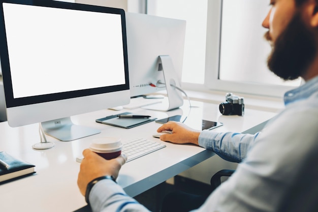 Cultivo barbudo sosteniendo una taza de bebida caliente y navegar por la computadora con pantalla en blanco mientras trabaja en la oficina