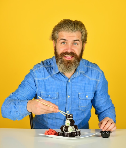 Cultura oriental. Hipster barbudo comiendo rollos. Entrega de comida japonesa. Rollos de sushi. Comida asiática. Comida para llevar sushi roll. El hombre come sushi en casa. Sushi y jengibre con palillos. Cocina japonesa.