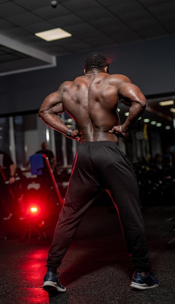 Culturista afroamericano muestra espalda fuerte sin camisa a la cámara. Atleta posa en el gimnasio.