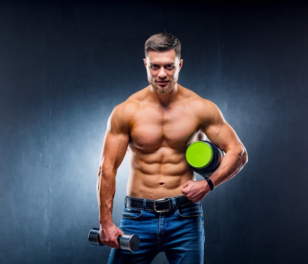 Culturista atleta masculino sostiene un frasco de nutrición deportiva y mancuernas en las manos. Posando para la cámara posa sobre fondo azul.