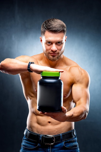 El culturista atleta masculino sostiene un frasco de nutrición deportiva en sus manos mostrando a la cámara poses sobre fondo azul.