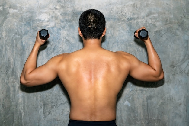 Culturista atlético hombre asiático posando con músculos de la espalda bumbbell, tríceps y latissimus en el fondo.