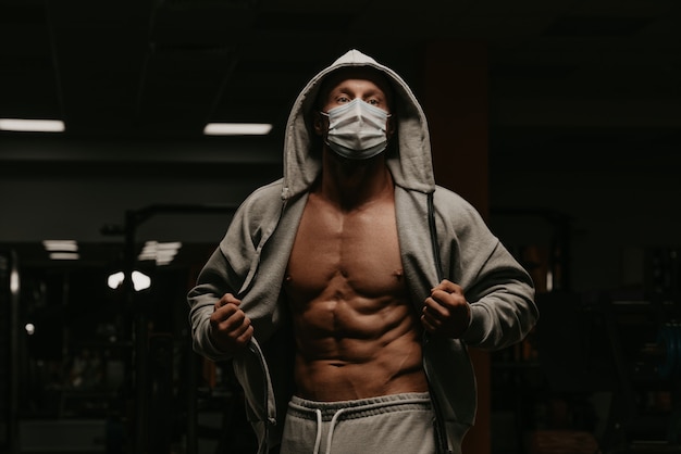 Un culturista con capucha y mascarilla para evitar la propagación del coronavirus abre una sudadera con capucha para demostrar su físico atlético. Un tipo deportivo con una mascarilla quirúrgica está posando después de un entrenamiento en un gimnasio.