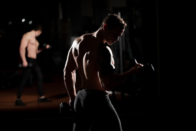 Culturista de chico atlético de poder guapo haciendo ejercicios con mancuernas. Cuerpo musculoso fitness sobre fondo oscuro.
