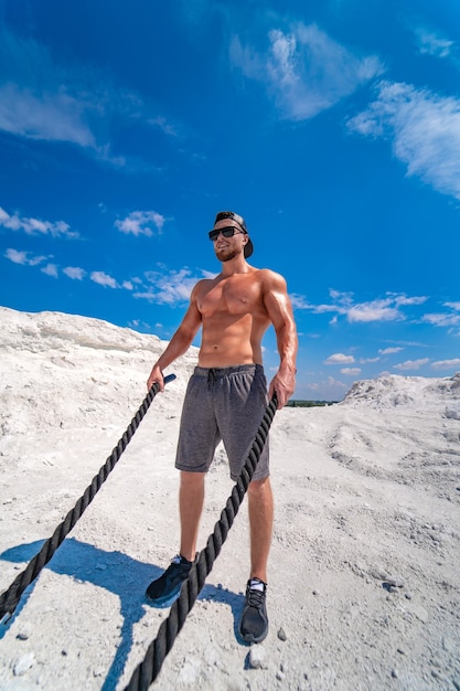 Culturista fuerte con una gorra y gafas de sol entrenando con cuerdas al aire libre. Entrenamiento pesado en una cantera. Paisaje blanco.