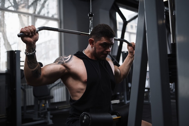 Culturista fuerte muscular que ejercita en la máquina del gimnasio del tirón del lat de la fila vertical