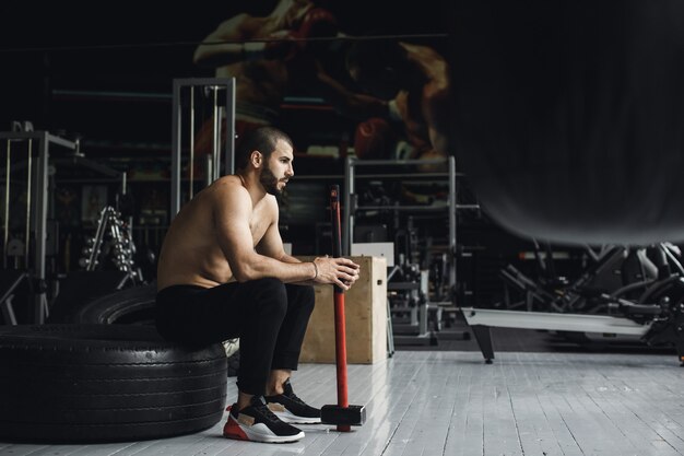 Culturista guapo con los ojos cerrados está descansando después de hacer ejercicios de mazo en el gimnasio. Foto de vista lateral de cuerpo entero. Foto de alta calidad