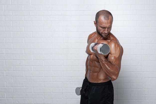 Foto culturista haciendo ejercicios para bíceps con pesas contra la pared de ladrillo.