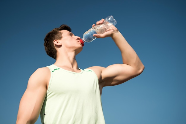 Culturista con manos musculosas músculos bíceps tríceps Deporte y concepto de fitness Salud y estilo de vida saludable Hombre bebiendo agua de la botella en un día soleado Atleta en camiseta verde en el cielo azul