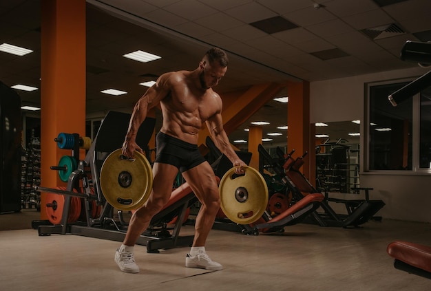 Un culturista en pantalones cortos camina por el gimnasio con dos placas de goma. Un deportista sin camisa está entrenando solo.