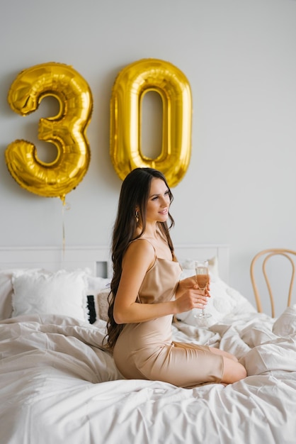 Una cumpleañera de pelo largo sentada en la cama por la mañana y sostiene una copa de champán en sus manos y sonríe celebrando su trigésimo cumpleaños