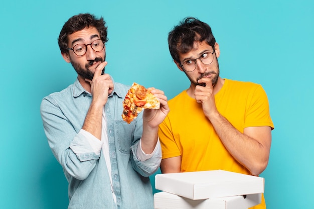 Cuople de dos amigos hispanos pensando en expresión y sosteniendo pizzas para llevar