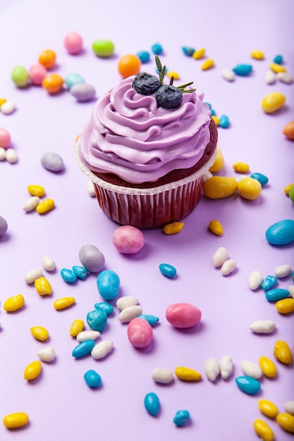 Cupcake de arándanos deliciosos dulces con crema y bayas colocados en medio de varios caramelos de grageas azucaradas multicolores sobre fondo lila