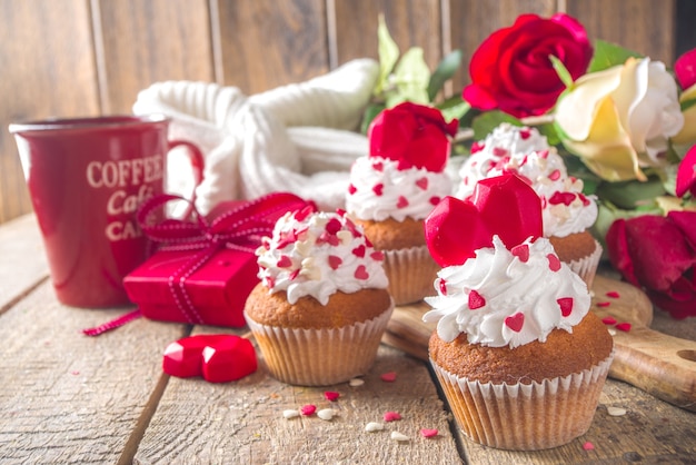 Cupcake com coração para dia dos namorados. sobremesa doce de dia dos namorados, cupcakes de baunilha com creme de baunilha e decoração de corações de açúcar vermelho para o dia dos namorados, mesa de madeira com buquê de flores de rosas