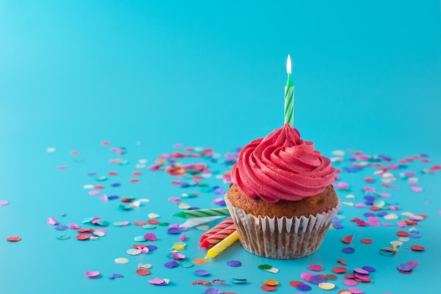 Cupcake de cumpleaños rosa con vela verde y confeti sobre fondo azul.