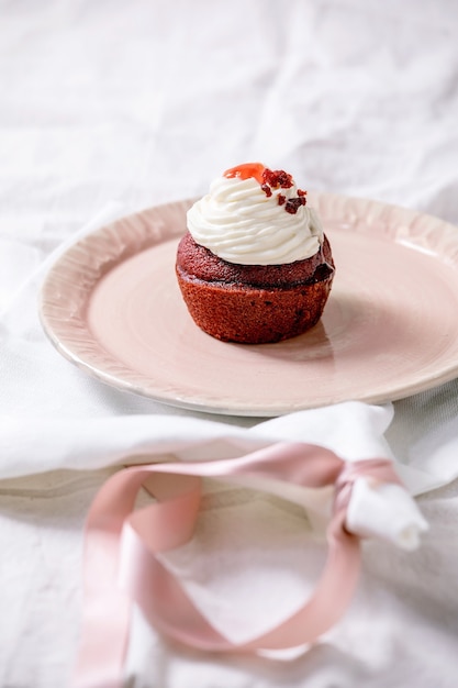 Cupcake de veludo vermelho caseiro com chantilly em prato de cerâmica rosa, guardanapo branco com fita em toalha de mesa de linho branco