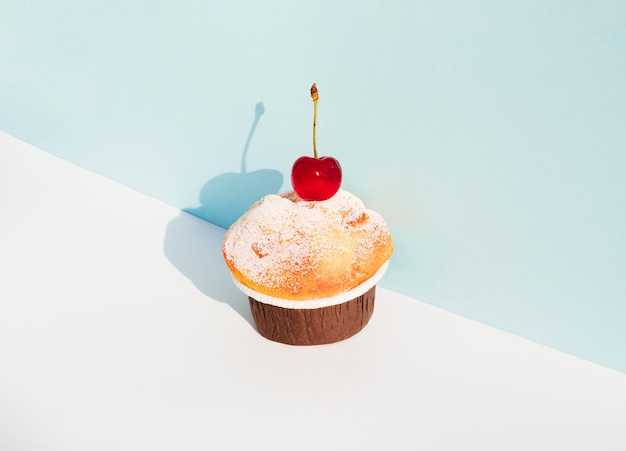 Cupcake mit Kirsche vor pastellblauem und weißem Hintergrund Sonniger Tagesschatten Minimales Essenskonzept