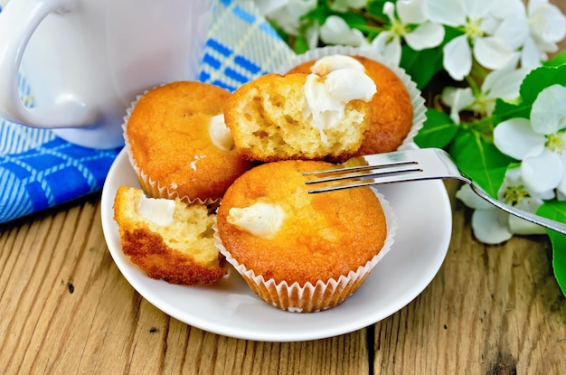 Cupcake mit süßer Sahne auf einem Teller, eine Tasse auf einer Serviette, ein Apfelblütenzweig, eine Gabel auf dem Hintergrund von Holzbrettern