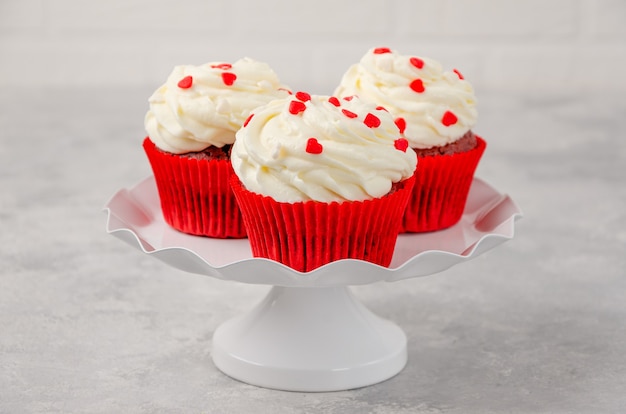 Los cupcakes de terciopelo rojo con glaseado de queso crema están decorados para el Día de San Valentín en un soporte blanco sobre un fondo gris. Copie el espacio.