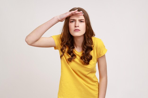 Curiosa adolescente de cabelos castanhos em camiseta casual, mantendo a palma sobre a cabeça e olhando atentamente para longe, parece com expectativa a longa distância. Tiro de estúdio interior isolado em fundo cinza.