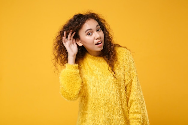 Curiosa joven afroamericana con suéter de piel posando aislada en el fondo de la pared naranja amarilla, retrato de estudio. Concepto de estilo de vida de las personas. Simulacros de espacio de copia. Trate de escucharlo con la mano cerca del oído.