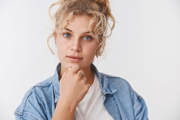 Curioso atractivo enfocado elegante millennial rubia escandinava de pelo rizado ojos azules frotándose la barbilla mirada intrigada escuchar interés de pie pensativo, pensando, de pie en la pared blanca