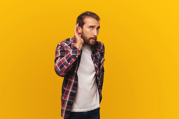 Curioso joven barbudo en camisa escucha chismes o intenta escuchar un secreto sobre fondo amarillo