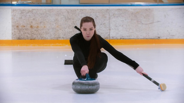 Curling entrenando a una mujer joven con el pelo largo en la pista de hielo sosteniendo una piedra de granito