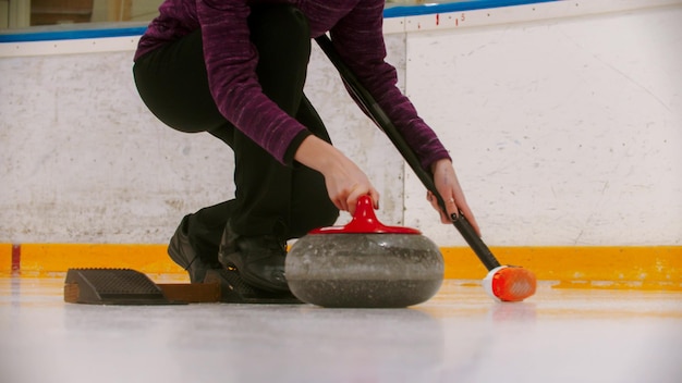 Foto curling treinando dentro de casa uma mulher prestes a empurrar o suporte no campo de gelo com pedra de granito