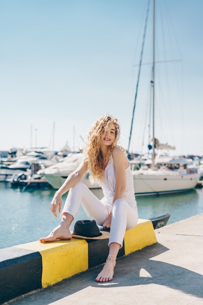 Curly Blonde mit einem Lächeln in einer entspannten Pose sitzt auf der Brüstung des Piers vor dem Hintergrund von Booten. barfuß