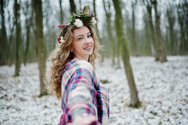 Curly linda garota loira com coroa de flores na manta quadriculada no bosque nevado em dia de inverno.