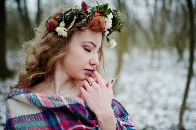 Curly linda garota loira com coroa de flores na manta quadriculada no bosque nevado em dia de inverno.