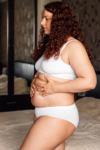 Curly Plus Size Frau in weißer Unterwäsche, die neben dem Bett im Schlafzimmer steht und den Bauch mit Dehnungsstreifen berührt Nahaufnahme Seitenansicht Fettverbrennung Behandlung von dickem Bauch Gewichtsverlust Programm Diät