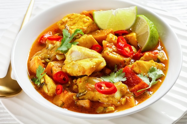 Curry brasileño de pescado y marisco Moqueca Baiana con pescado blanco, leche de coco, varias verduras, gajos de lima y cilantro fresco en un tazón blanco cerrado