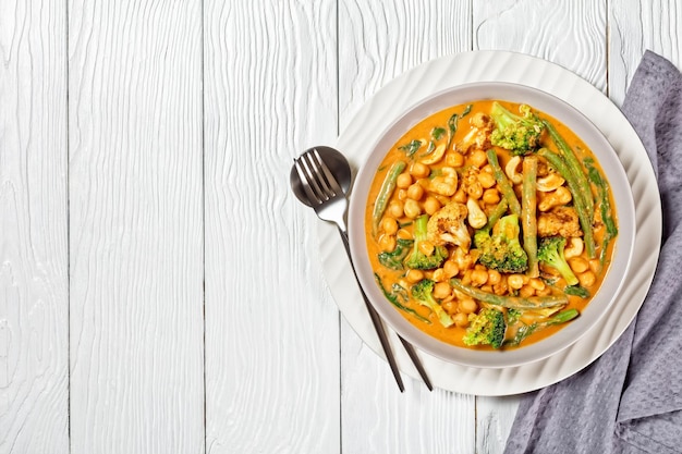 Curry de coco vegetariano con garbanzos, judías verdes, coliflor y floretes de brócoli en un recipiente sobre una mesa de madera blanca, plano, espacio libre, cocina india