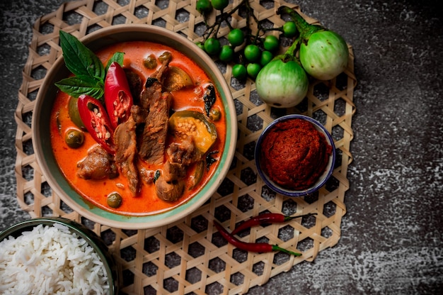 Curry tailandés picante con carne de cerdo que se sirve con arroz y decora con ingredientes vegetales a base de hierbas como el chile y la berenjena sobre fondo rústico