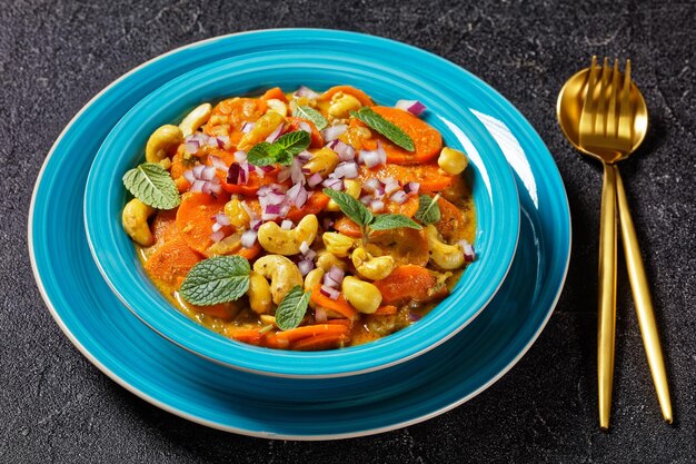 Curry de zanahoria con anacardos y pasas en un recipiente azul sobre una mesa de hormigón, vista horizontal desde arriba, primer plano, cocina india