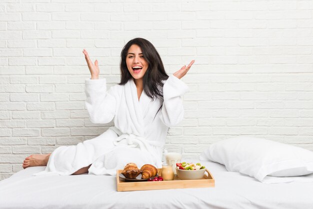 Foto curvilínea jovem tomando um café da manhã na cama recebendo uma agradável surpresa, animada e levantando as mãos.