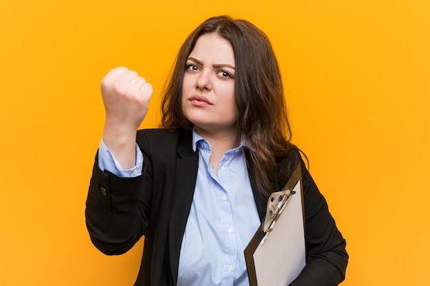 Curvilíneas novas, mais tamanho mulher de negócios holdingclipboard mostrando o punho, expressão facial agressiva.