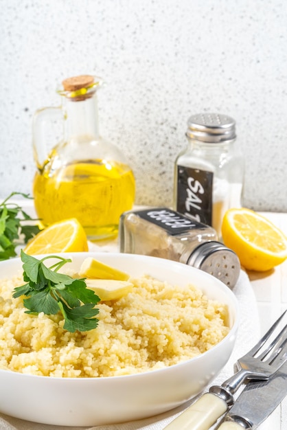 Cuscús cocido con cilantro o mantequilla de perejil, aceite de oliva, limón en un tazón de cerámica blanca en la mesa blanca de la cocina, espacio para copiar